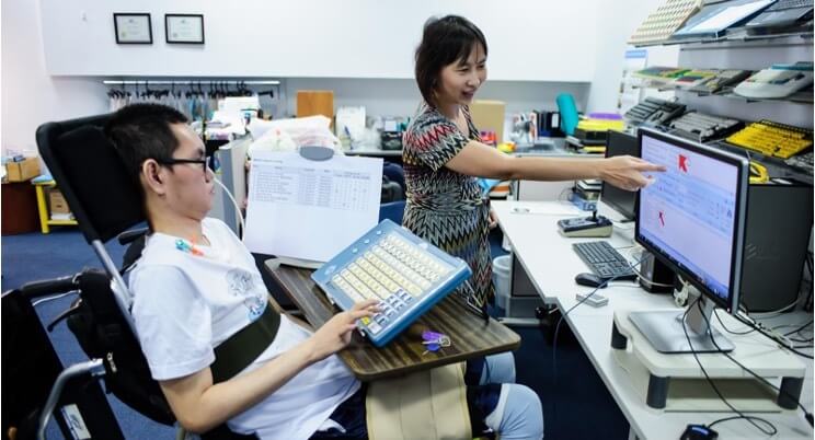 one person in a wheelchair using a special keyboard with big keys with one hand while another person is showing the result on screen, the cursor is a big red arrow