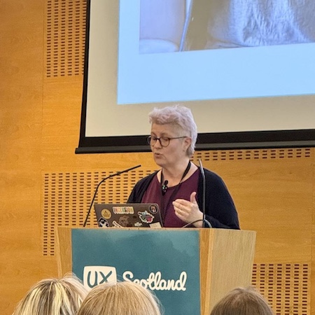 Me, Stephanie, standing behind a wood lectern with a poster of the UX Scotland logo. I'm looking at my notes on my laptop